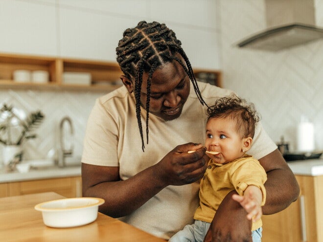 Dad feeding his baby girl, sitting on his lap.